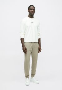 Cămașă albă cu mâneci lungi și logo-ul "EA7", asortată cu pantaloni sport bej și adidași albi. Design simplu, cu manșete la mâneci și pantaloni.