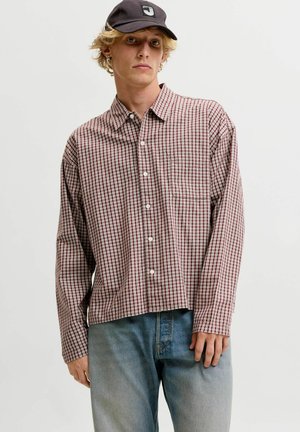 Jeune homme portant une chemise à carreaux rouge et blanc boutonnée, un jean bleu clair et une casquette de baseball noire sur un fond uni.