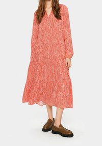 Červené midi šaty se vzorem, dlouhými rukávy, V-výstřihem a tiered volánovým lemem. Nosit s hnědými mokasíny a texturovanou tkaninou.