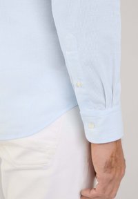 Camisa a rayas azul claro con mangas largas, que presenta puños abotonados y una textura suave. Pantalones blancos son parcialmente visibles debajo de la camisa.