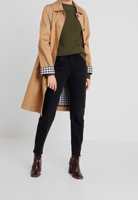 Beige Trenchcoat mit Futter im schwarz-weißen Karomuster, olivgrüner Pullover, schwarze Hose und braune Stiefel mit Absatz.