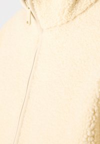 Giacca in pile color crema con una superficie morbida e strutturata. Presenta una cerniera centrale e un colletto alto per un calore extra.