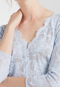 Ljusblå spetsblus med V-ringning och scallopade kanter samt 3/4-långa ärmar. Texturerat blommönster över hela plagget.