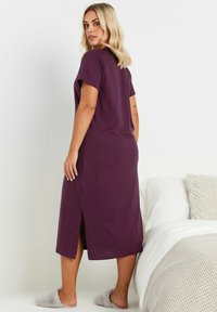 Lila, kortärmad nattklänning med slits på sidan, tillverkad av mjukt tyg. Bärs med grå, fluffiga tofflor, mot en vit sängöverkast och en strukturerad pläd.