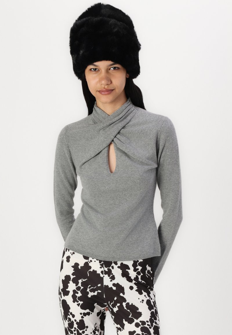 Graues Langarmoberteil mit drapiertem Ausschnitt und Schlitzdetail, kombiniert mit einer Hose im schwarz-weißen Kuhfleckenmuster. Schwarze Kunstfellmütze.