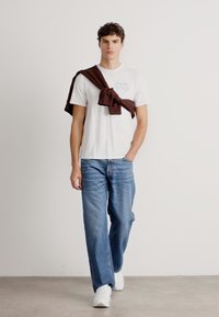 Un tricou alb cu text imprimat, asortat cu blugi ușori albaștri și adidași albi. O jachetă maro este drapată pe umeri.