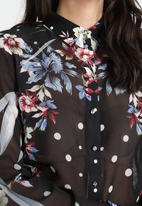 Sort gennemsigtig bluse med blomstermønster i rød, blå og hvid, med prikker og en knaplukning foran med dekorative knapper.