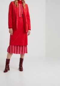 Rode corduroy blazer over een roze en rode gestreepte jurk, gecombineerd met glanzende bordeauxrode enkellaarsjes. Soepele texturen en gedurfde kleurcontrasten.