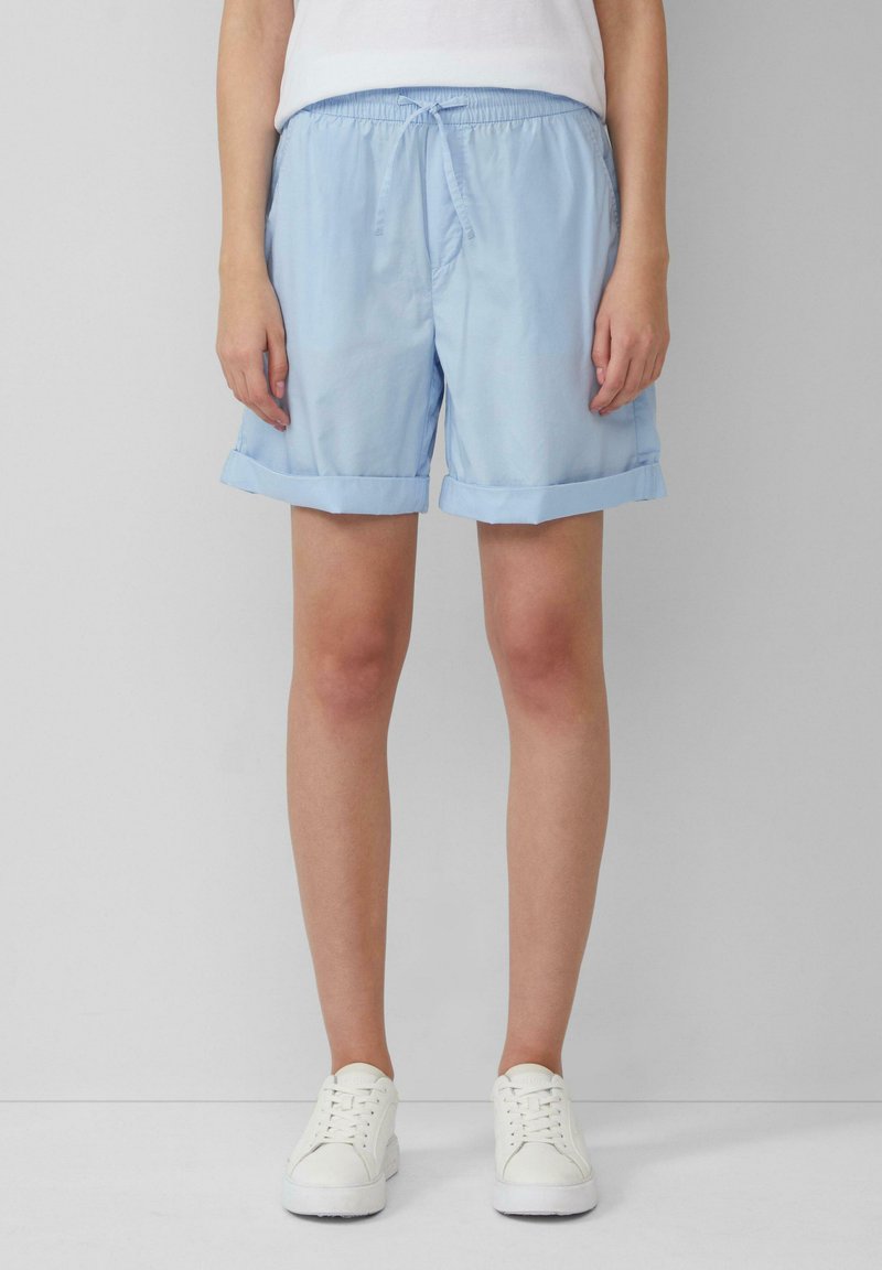 Shorts bleu clair, avec une taille élastique et un cordon de serrage, présentant des revers et un tissu lisse et léger. Associés à des baskets blanches.