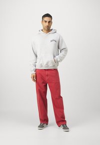 Gray sweatshirt with a front pocket and logo, paired with loose red pants and gray sneakers, standing against a plain white background.