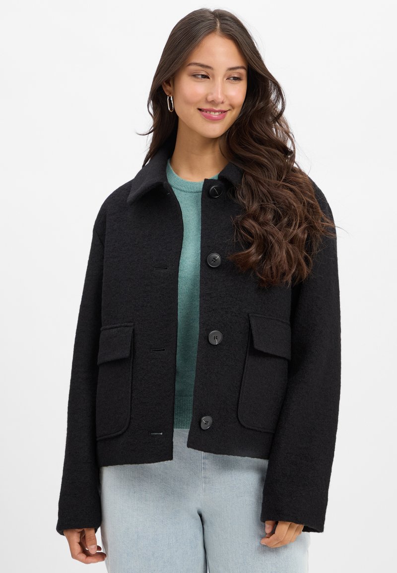 Black wool-blend cropped jacket with a collar, button closure, two large front pockets, and a textured surface. Paired with a green sweater.