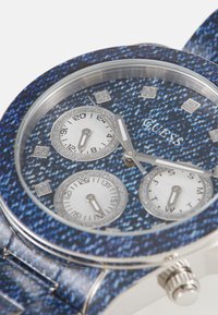 Reloj Guess con patrón de denim azul, tres sub-diales, marcadores en tono plateado y hardware de acero inoxidable; correa y bisel de tela texturizada.