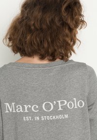 Sudadera de algodón gris con un escote ancho y texto impreso en blanco: "Marc O'Polo EST. EN ESTOCOLMO" en la parte trasera.