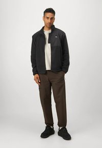 Giacca in pile nera con zip e collo alto, maglietta grigio chiaro sotto, pantaloni marroni e scarpe da ginnastica nere. Design semplice e funzionale.