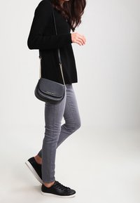 Ristilaukku tekstuuripinnasta harmaassa materiaalissa, ketjuhihnalla ja vetoketjusuljennalla. Yhdistetty harmaisiin farkkuihin ja mustiin lenkkareihin.