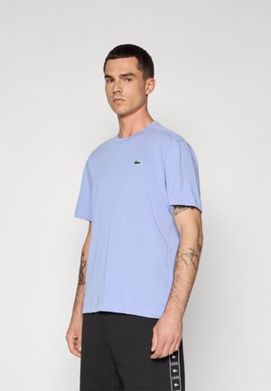 Világoskék, puha pamutból készült póló, kerek nyakkivágással és rövid ujjakkal. A mellkason egy kis zöld Lacoste logó található.
