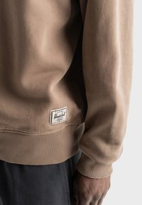 Bruine sweatshirt met ribbels aan de manchetten en onderkant. Beschikt over een rechthoekig wit label met zwarte tekst dat aan de onderste linkerkant is genaaid. Zachte textuur.