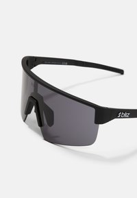 Gafas de sol deportivas negras con una gran lente gris curvada, un marco resistente y un soporte de puente nasal único. Logo visible en el brazo lateral. Diseño minimalista.