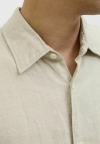 Camisa de lino beige claro con cuello puntiagudo, presenta una tapeta de botones y un acabado de tela texturizada. Vista en primer plano del escote.