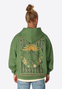 Grünes, übergroßes Hoodie mit einem Grafikdruck auf dem Rücken, der eine Sonne, Berge und stilisierte Schrift in Gelb und Beige zeigt. Weicher Stoff.