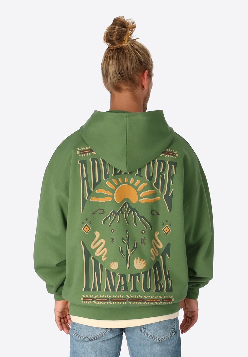 Grünes, übergroßes Hoodie mit einem Grafikdruck auf dem Rücken, der eine Sonne, Berge und stilisierte Schrift in Gelb und Beige zeigt. Weicher Stoff.