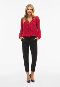 Blusa rossa con dettagli di volant, scollatura a V e maniche lunghe abbinata a pantaloni neri aderenti e scarpe con tacco nere.