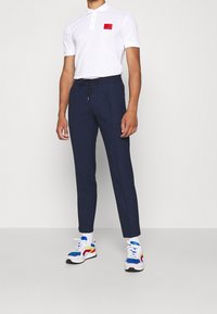 Polo blanc avec un écusson logo rouge, pantalon bleu marine à cordon de serrage, et baskets multicolores avec des accents bleus, jaunes et rouges.
