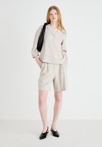 Camicia button-up in lino beige con colletto, abbinata a pantaloni corti coordinati. Il modello indossa tacchi slingback neri e porta una borsa tote nera.