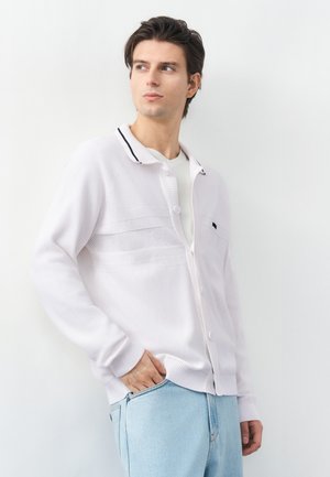 Noor mees tume juustega, seljas valge nööbitav kardigan ja helesinised teksad, seisab valge seina ääres ja vaatab vasakule.