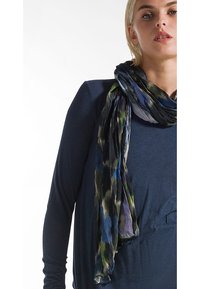 Sciarpa leggera e multicolore con un mix di schemi astratti blu e verdi, caratterizzata da una consistenza morbida e fluida. Stile di avvolgimento mostrato.