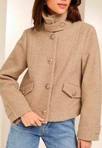 Giacca in misto lana beige con colletto alto, bottoni dorati, tasche frontali e maniche leggermente accorciate. Texture morbida con un design strutturato.