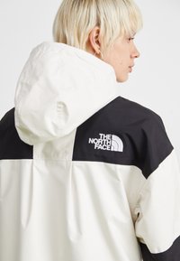 Veste à capuche blanche et noire avec un logo au dos, fabriquée en matériau lisse, avec une coupe décontractée et un blocage de couleurs contrastées.