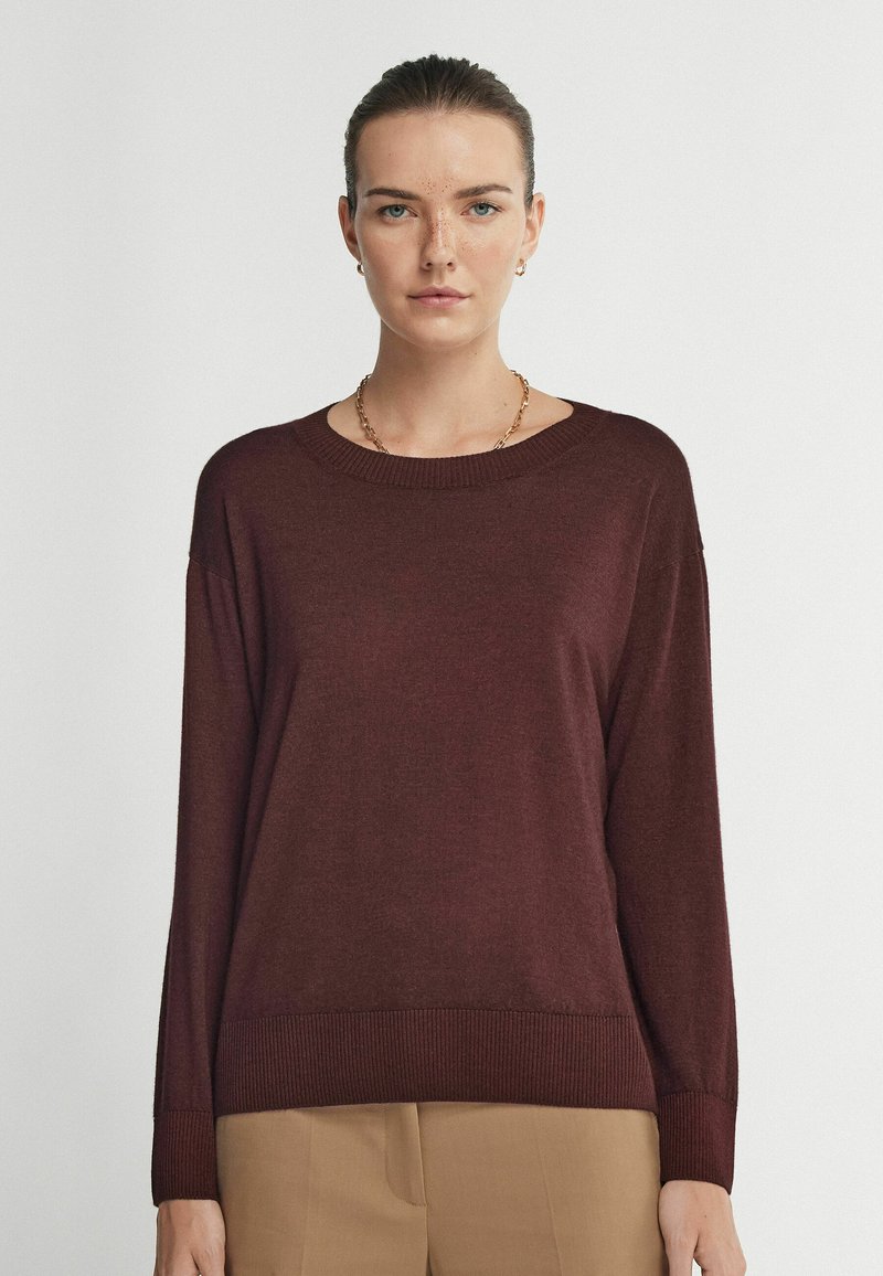 Maroon stickad tröja med rund halsringning, långa ärmar, ribbad nederkant och en avslappnad passform. Slät textur utan synliga mönster eller detaljer.