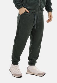 Pantaloni jogger in pile di un verde scuro, con vita elasticizzata e cordino regolabile; indossati con sneakers di colore chiaro con accenti grigi.