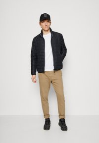 Zwarte gewatteerde jas met een hoge kraag, wit T-shirt, beige chino's en zwarte sneakers, aangevuld met een zwarte cap met een logo.