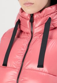 Giacca puffa rosa imbottita con colletto alto, zip nera e cordino. Texture lucida con sezioni a quadri orizzontali.