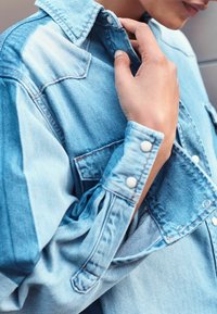 Camicia in denim azzurro chiaro con colletto a punta, chiusura a bottoni e tasche sul petto. Presenta una texture variegata e dettagli di cucitura visibili.