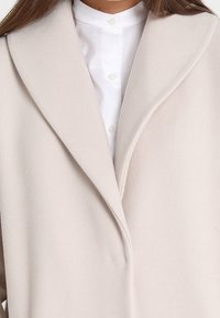 Béžový kabát s měkkou texturou, s velkým šálovým límcem a jedním zapínáním na knoflík, nošený přes bílou košili na knoflíky.