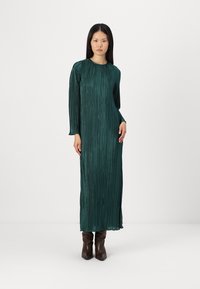 Μακριά πλεκτή φόρεμα σε σκούρο πράσινο, με μακριά μανίκια και στρογγυλή λαιμόκοψη. Φορεμένο με σκούρα καφέ μπότες ως το γόνατο. Λείος χαρακτήρας.