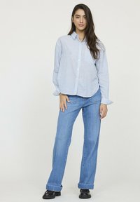 Chemise à rayures bleues et blanches à manches longues, avec un ajustement décontracté, assortie à un jean droit bleu clair. Des chaussures noires complètent le look.