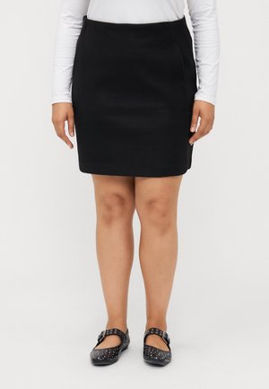 Femme portant une jupe noire longueur genou, un haut blanc à manches longues et des chaussures Mary Jane noires cloutées, debout devant un fond blanc.