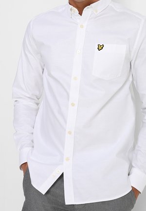 Chemise blanche boutonnée à manches longues avec une poche poitrine arborant un petit logo d'oiseau jaune et noir, portée avec un pantalon gris.