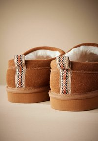 Brune ruskinds slippers med en blød hvid foring, der har et dekorativt vævet bånd i jordfarver af orange, sort og creme.