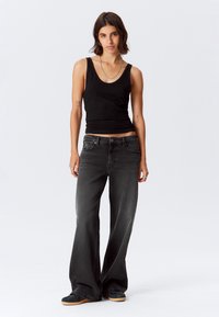 Donna con capelli corti castani, che indossa una canotta nera, jeans larghi grigio scuro, scarpe da ginnastica nere e gioielli in argento, in piedi davanti a uno sfondo bianco.