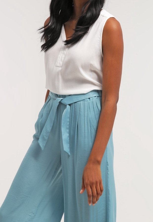 Blouse blanche sans manches avec un plastron sur le devant. Pantalons à taille haute, larges et de couleur bleu clair avec une ceinture à nouer et une texture douce.