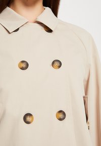 Trench beige con un cuello clásico, que cuenta con tres grandes botones contrastantes con un patrón veteado y bolsillos laterales. Textura de tela suave.
