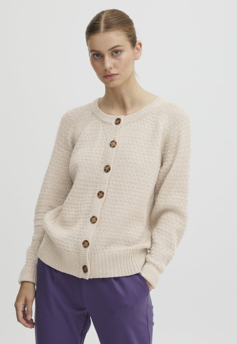 Beige pletený cardigan s texturovaným vzorem, kulatým výstřihem a dřevěnými knoflíky na přední straně; kombinovaný s fialovými kalhotami.