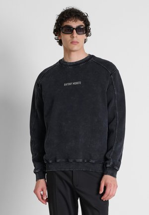 Jonge man met krullend haar die een zwarte oversized trui draagt met de tekst "Antony Morato" en grote donkere zonnebril, staand tegen een effen achtergrond.
