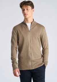 Jeune homme portant un pull à fermeture éclair marron sur une chemise blanche, debout avec une main dans la poche contre un fond clair uni.