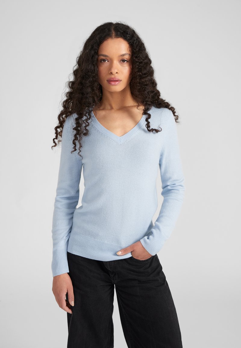 Pull léger bleu clair en tricot doux, avec un col en V, des manches longues et une coupe ajustée. Associé à un pantalon large noir.